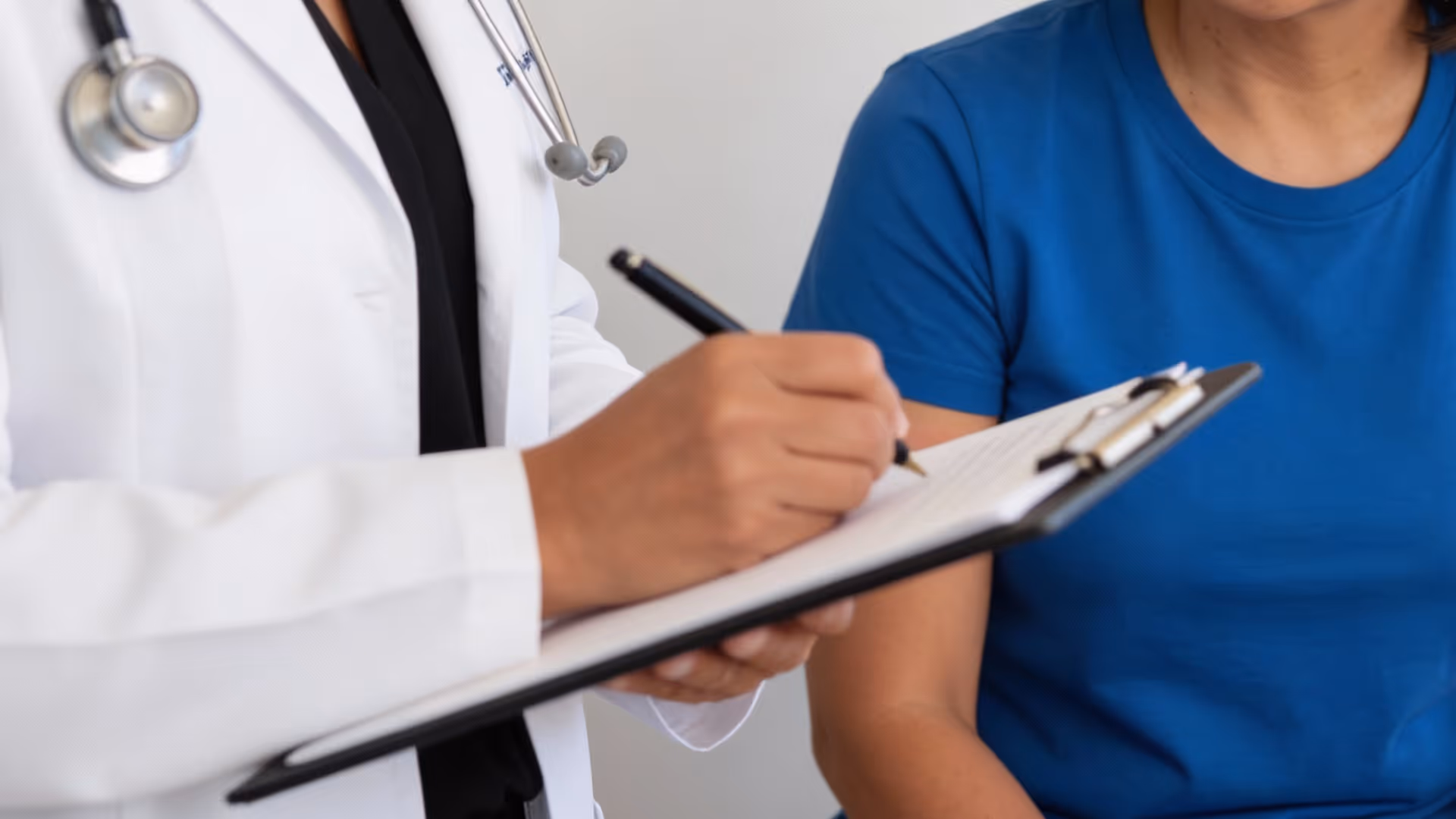 Médico escribe en una tabla con un bolígrafo mientras un paciente en camiseta azul espera.