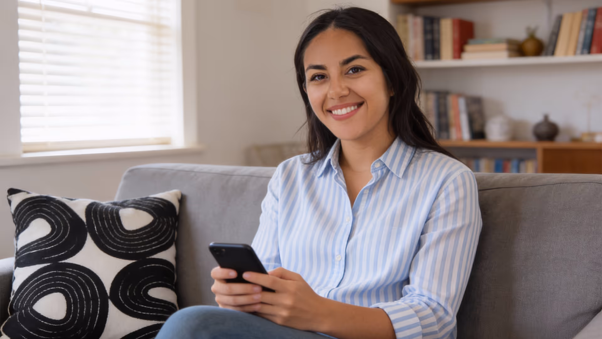Mujer sonriente con camisa a rayas azules, sentada en sofá gris con almohada, sosteniendo un teléfono móvil.