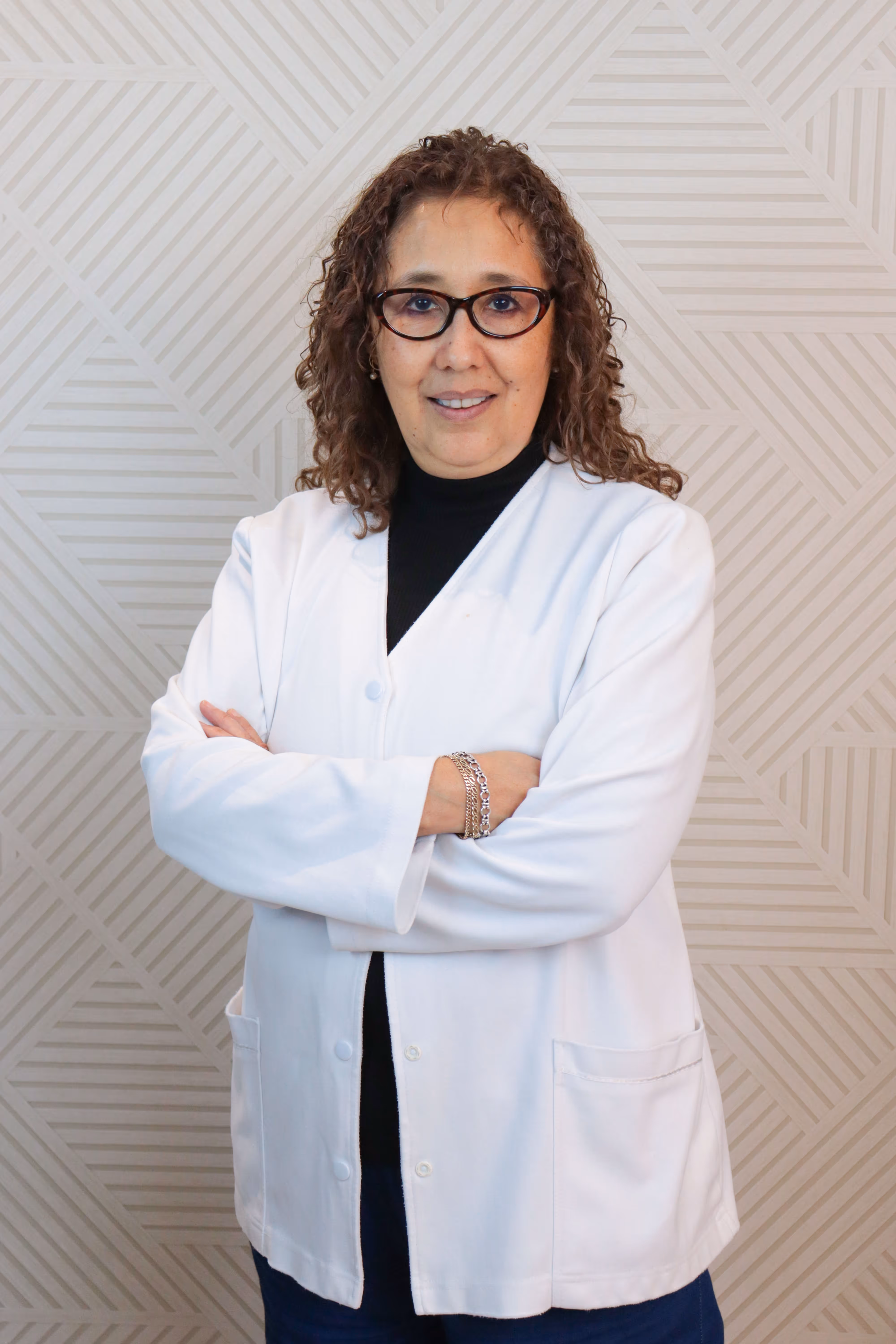 Mujer con cabello rizado y gafas, con bata blanca y camisilla negra, posando con brazos cruzados frente a pared con patrón geométrico.