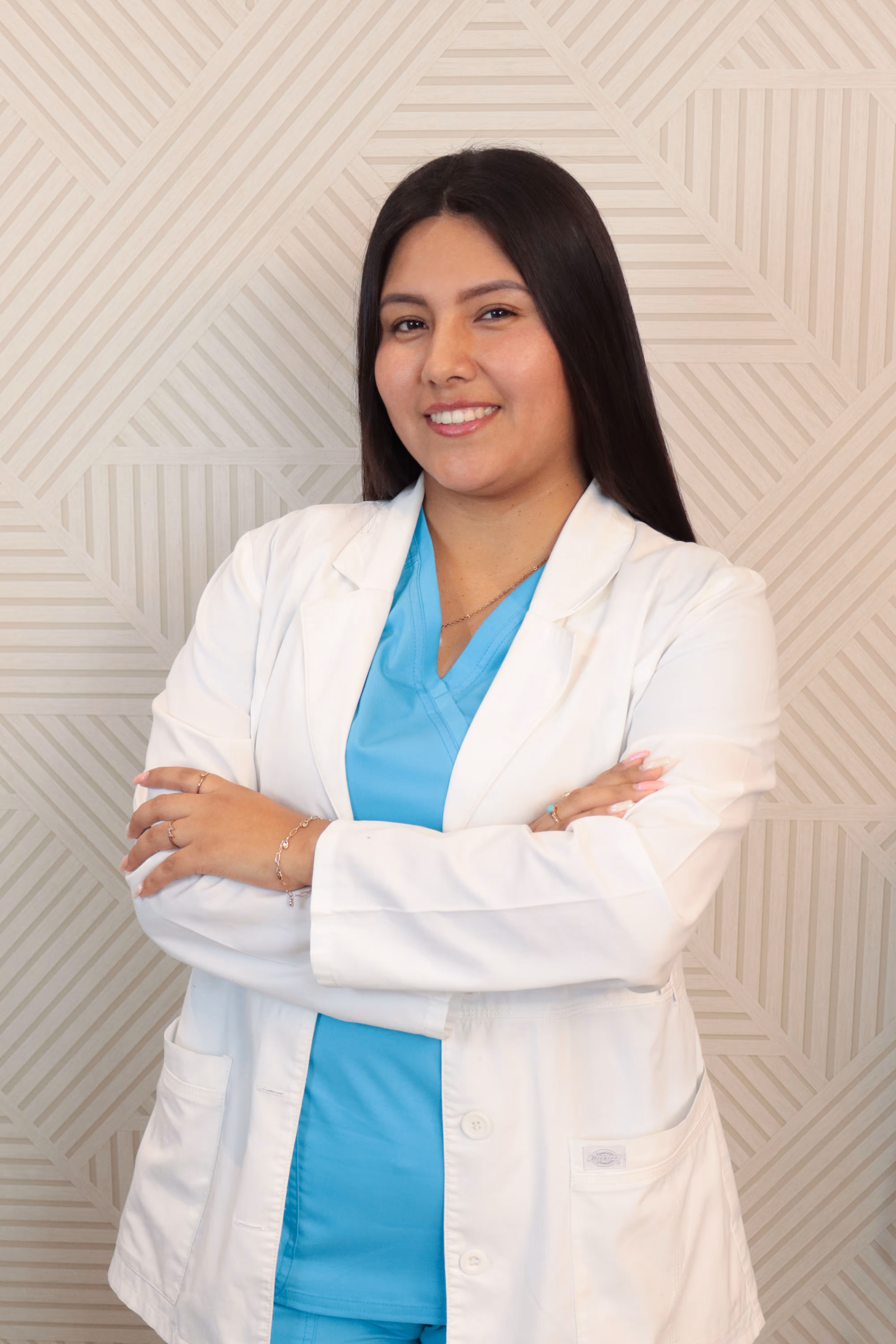 Mujer joven con bata blanca y uniforme médico azul, sonriendo y con los brazos cruzados frente a un fondo con patrón geométrico.