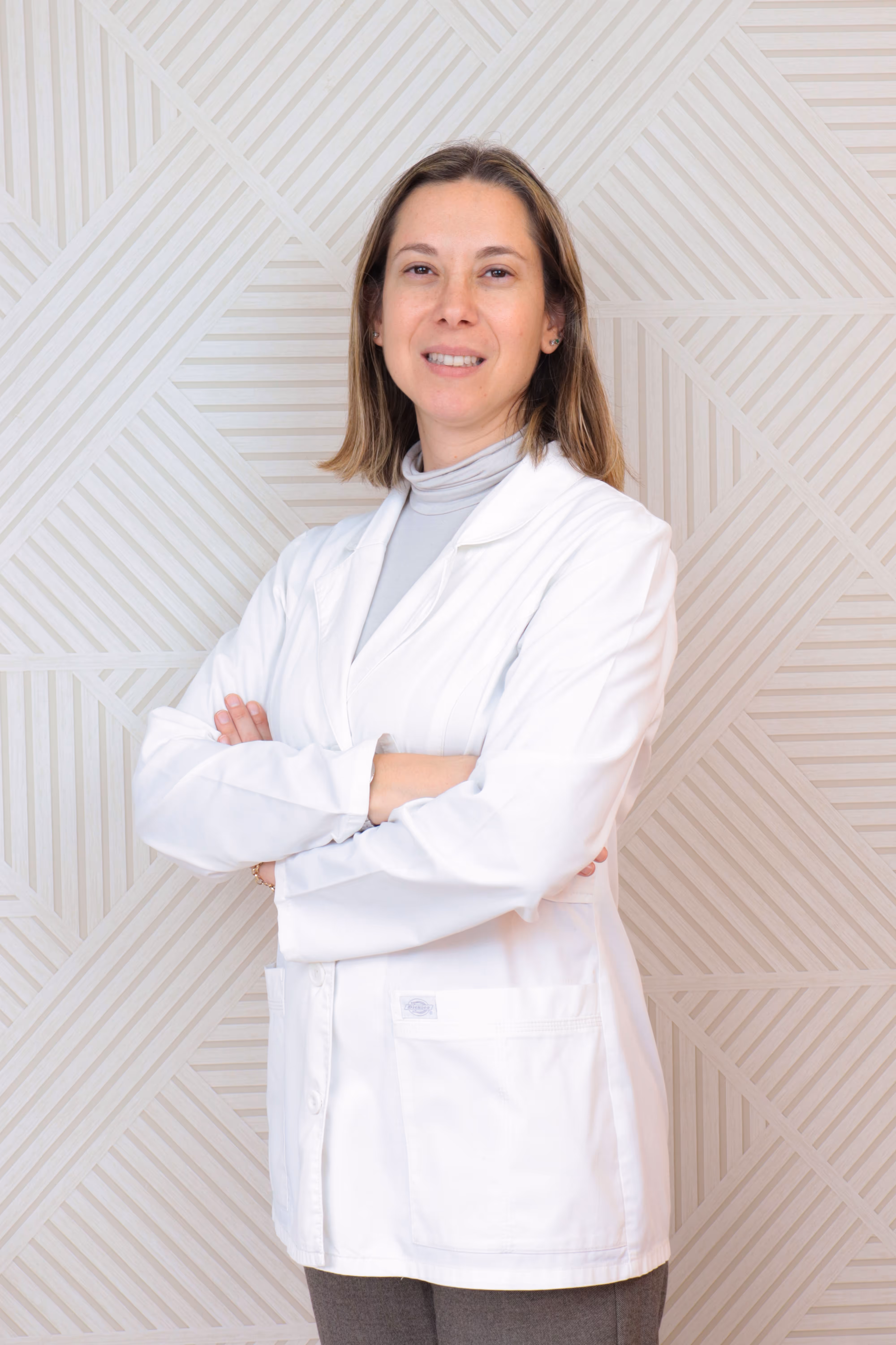 Mujer con bata blanca cruzada de brazos, sonriente, frente a una pared con patrón geométrico claro.