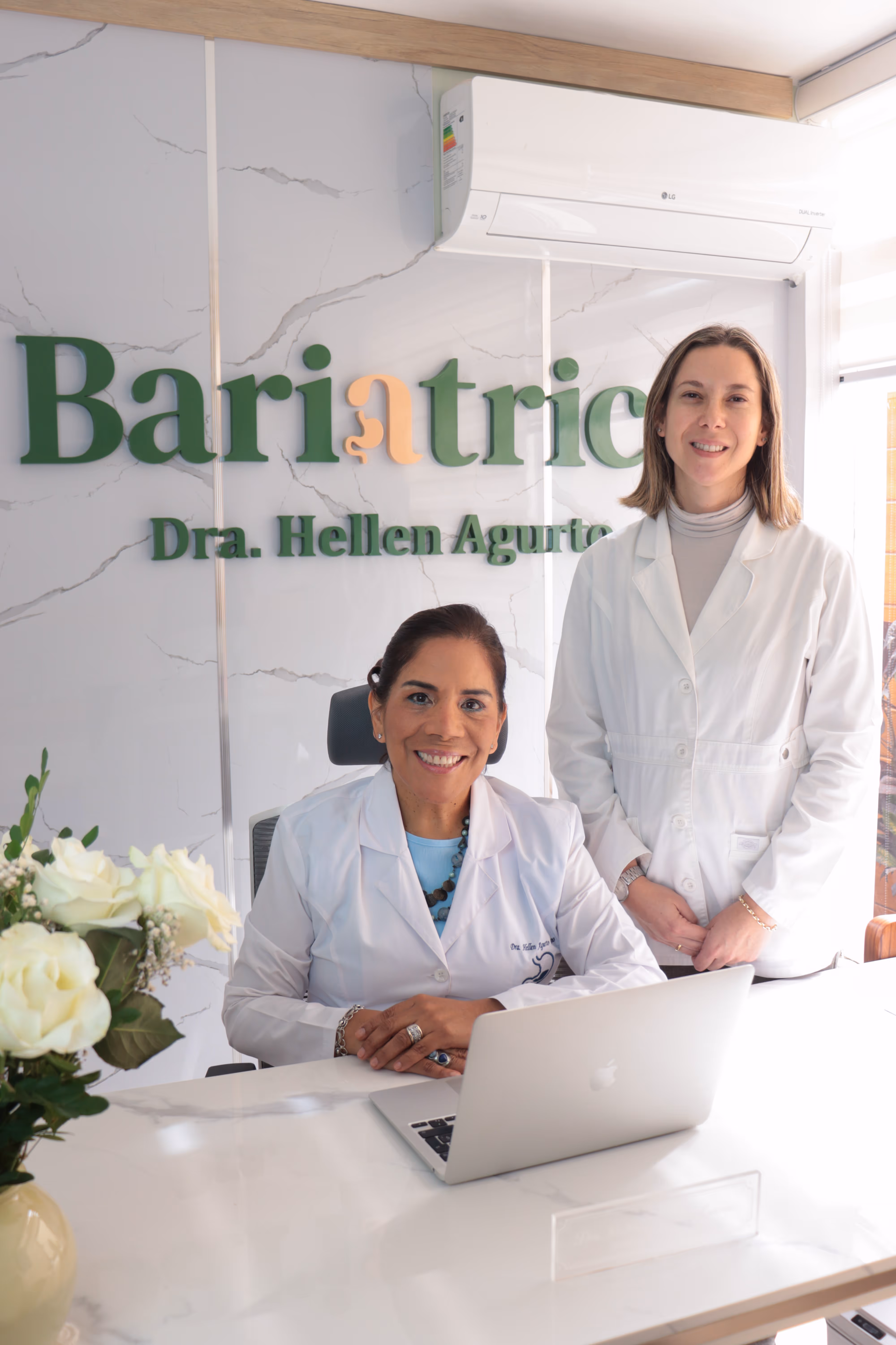 Dos doctoras en bata blanca en una consulta médica de bariatría, una sentada frente a una laptop y otra de pie.