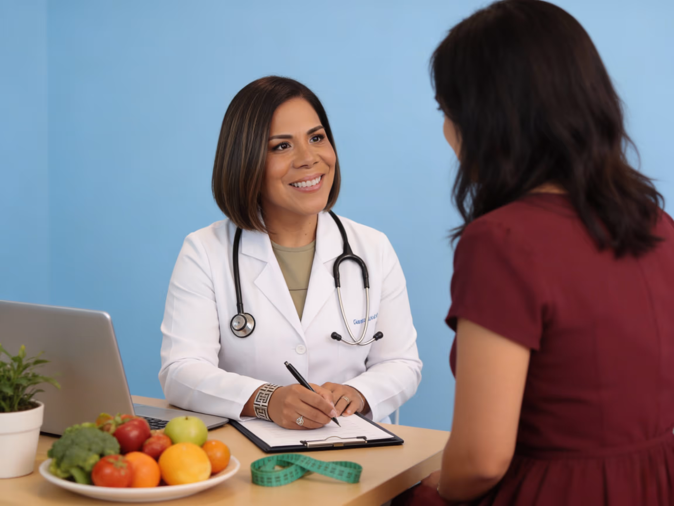 Doctora sonriente con estetoscopio en consulta hablando con una paciente frente a una mesa con frutas y una cinta métrica.